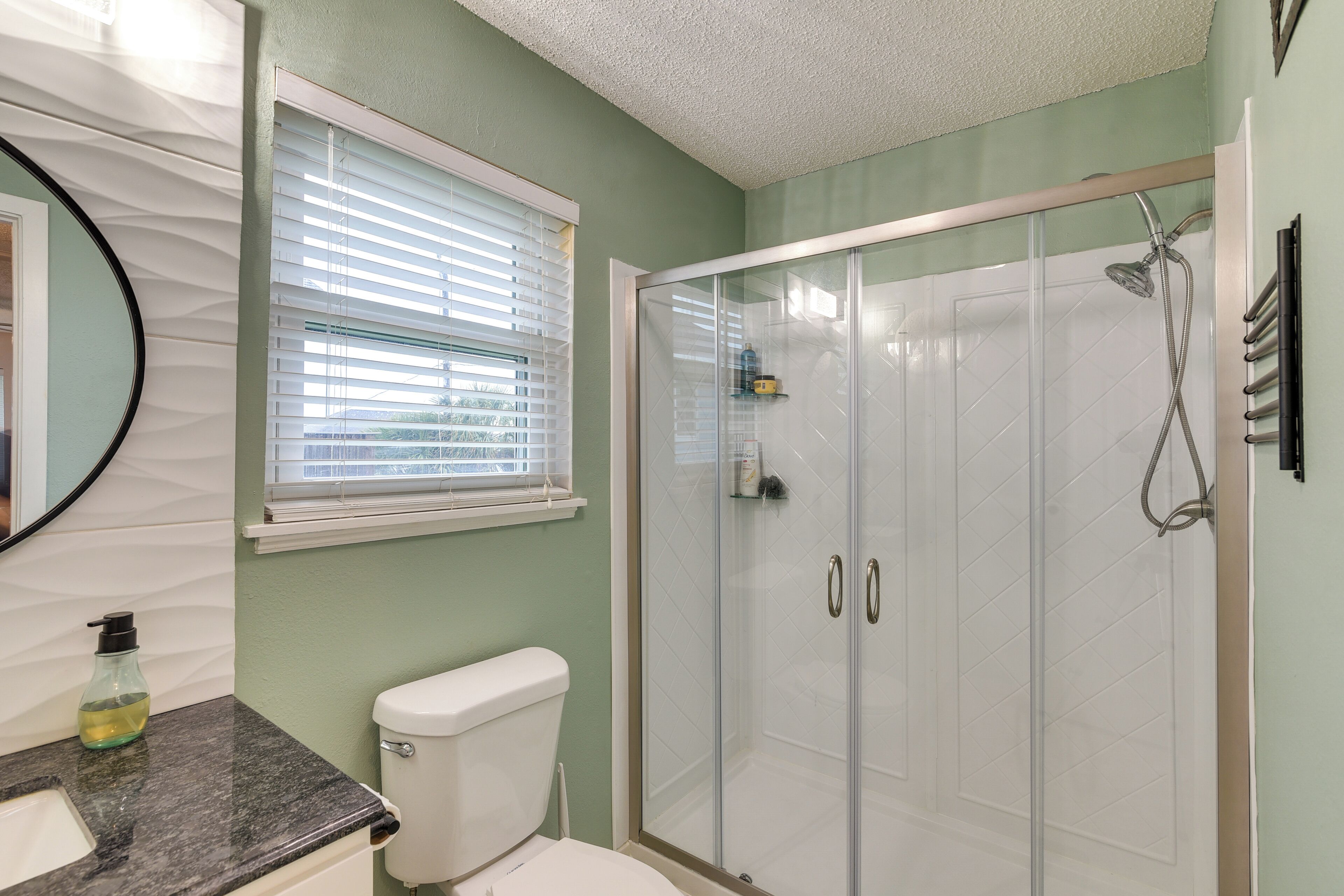 A bathroom with a toilet, a sink, a shower with glass doors, and a window with blinds.