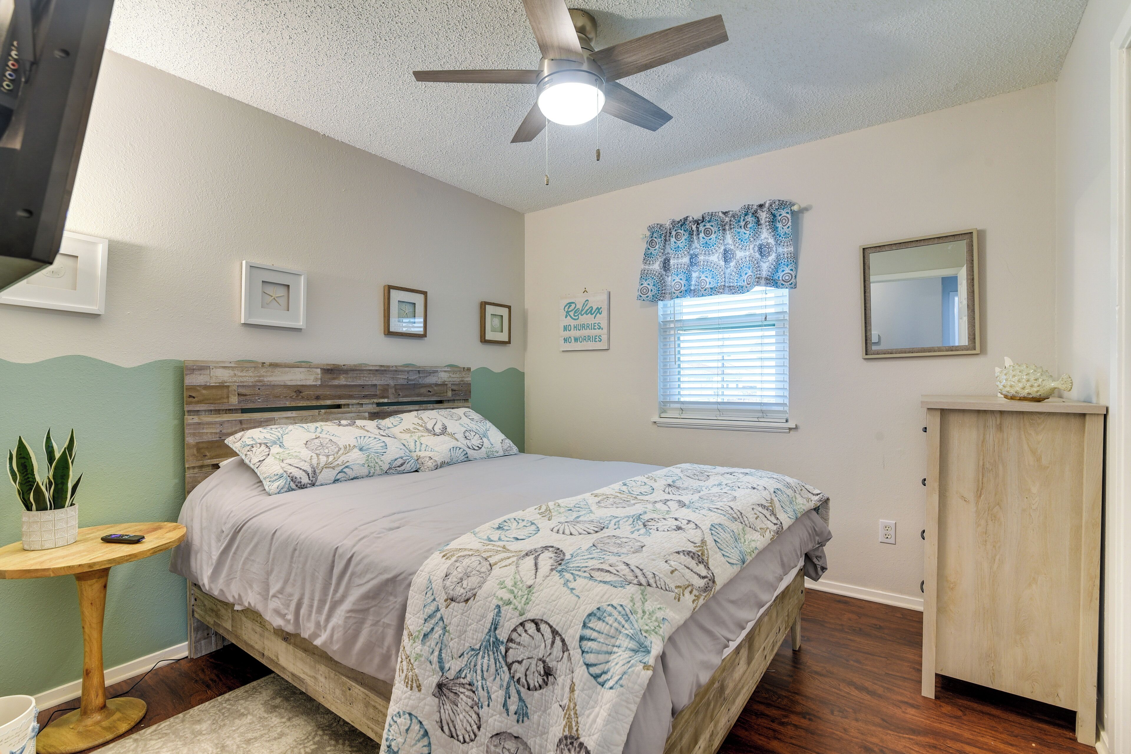 Une chambre à coucher comprenant un lit, un ventilateur de plafond, une fenêtre avec des stores, une petite table avec une plante et une armoire en bois.