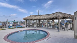 A swimming pool with a brick border, a shaded area with benches, and a fenced parking lot in the background.