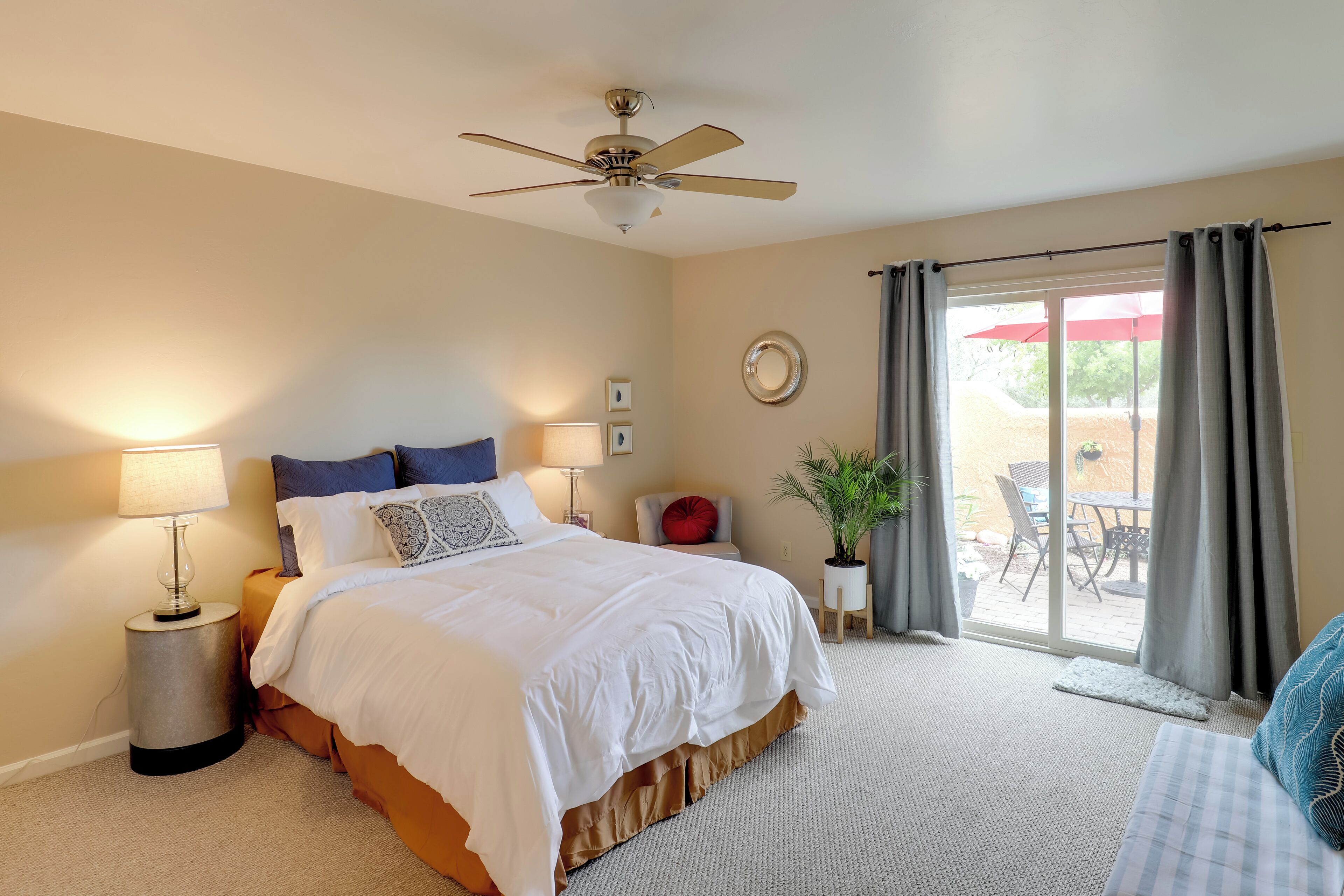 A bedroom with a bed, bedside lamps, a ceiling fan, a view of the patio through a sliding door, and a potted plant.