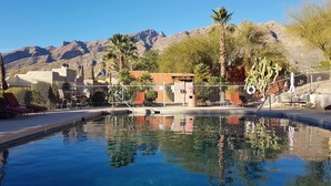 A swimming pool with clear water reflecting surrounding trees and buildings, a fenced area with lounge chairs, and mountains in the background.