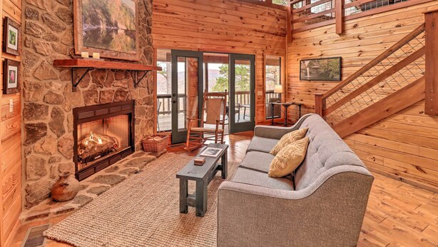 A cozy log cabin interior with a stone fireplace, a gray sofa, and a wooden staircase. - Piney Bluff Retreat With Mountain Views! (Melbourne)