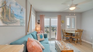 A living room with a blue sofa, a wooden coffee table, a dining table with chairs, a ceiling fan, and a painting of a beach scene. - Resort Perks: Gulf Shores Beach-inspired Condo! (Gulf Shores)