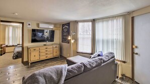 A living room with a sofa, a TV, a wooden cabinet, and a large window with blinds.