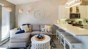 Uma sala de estar moderna com um sofá seccional cinza, uma mesa de centro redonda e uma área de cozinha com armários brancos e micro-ondas embutido.