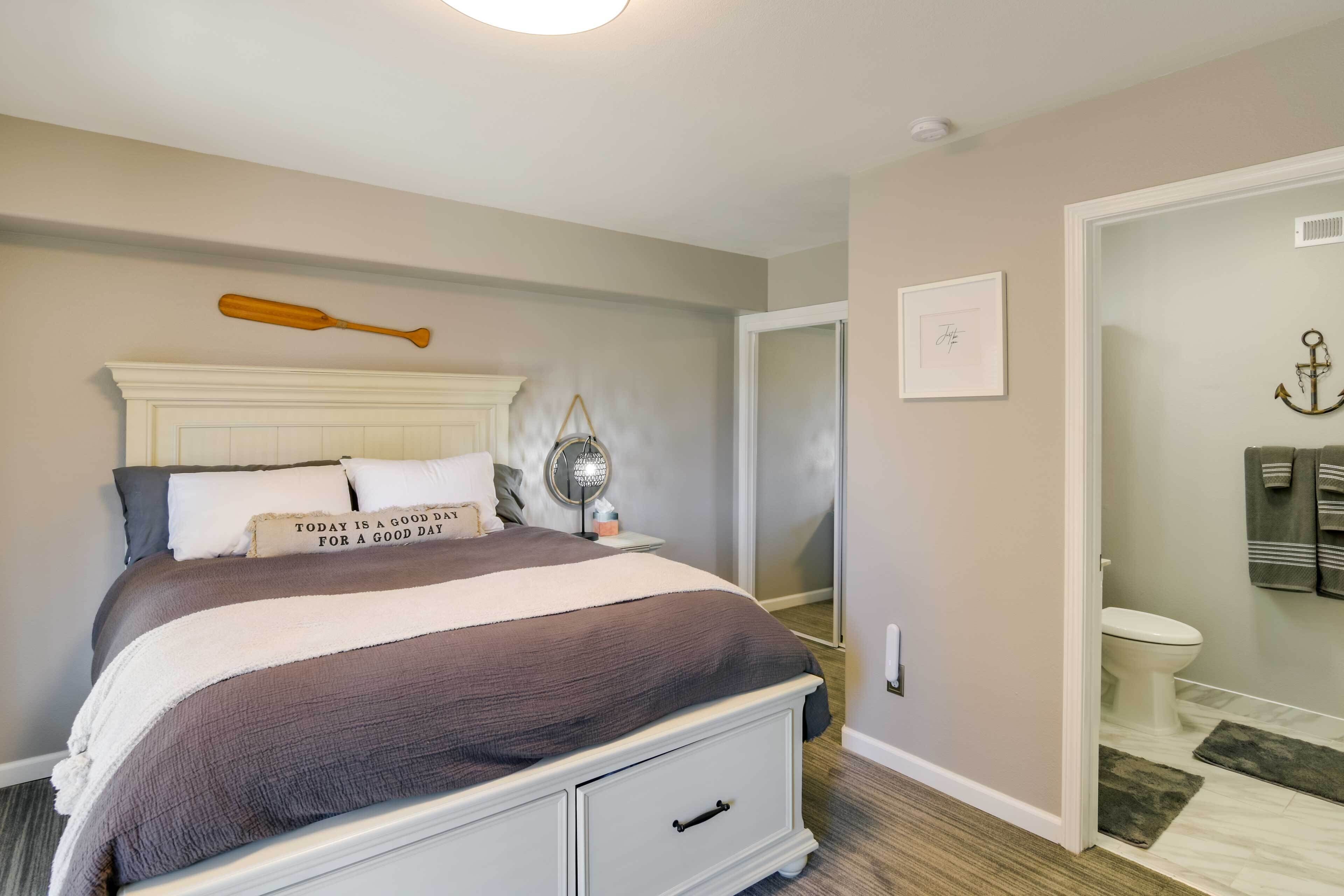 A bedroom with a bed, a bedside table, a nightstand, a mirror, a door, and a bathroom visible through an open door.