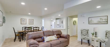 A spacious living room with a brown leather sofa, a dining table with chairs, and a kitchen area in the background.