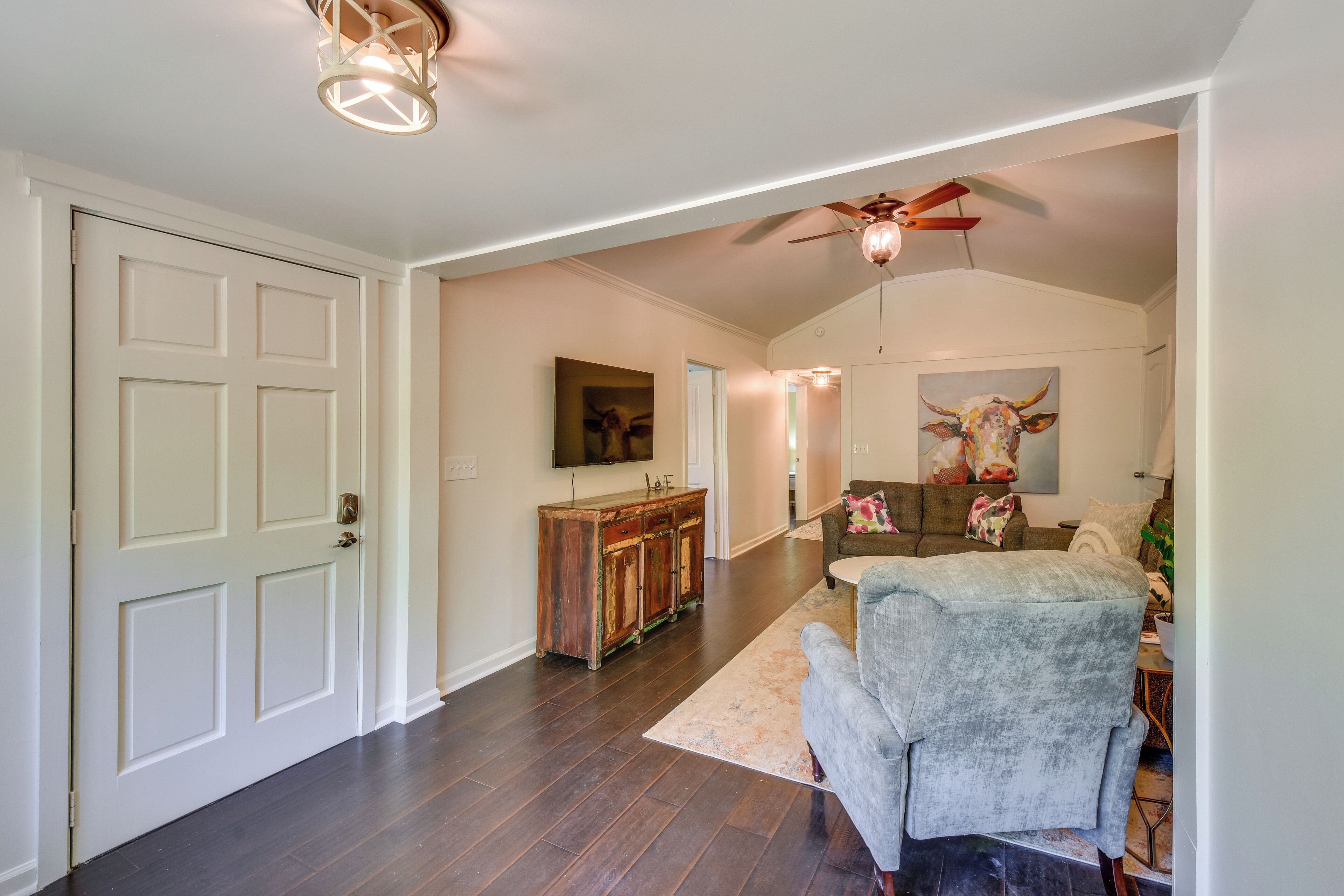 A spacious living room with a high ceiling fan, a wooden cabinet, a gray armchair, and a large painting of a bull.