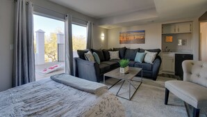 En moderne stue med sofa, sofabord og seng. Der er et tekøkken med skabe og vask.