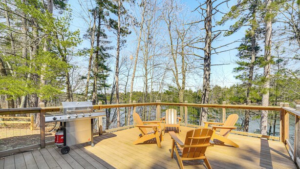Eine Holzterrasse mit Grill, Adirondack-Stühlen und Blick auf Bäume und Wasser.