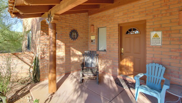 A covered porch with a wooden bench and a blue Adirondack chair, a brick wall, and a door with a caution sign. - Tucson Home - Hiking Trail Access On-site! (Tucson)