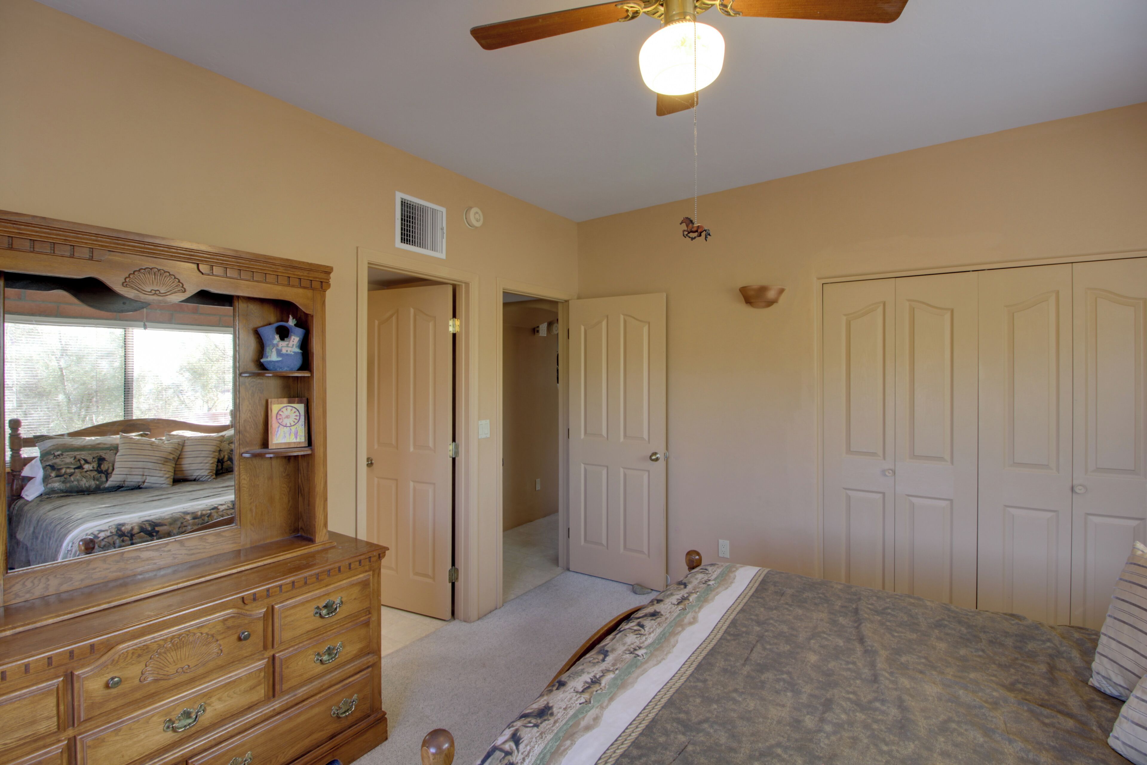 A bedroom with a wooden dresser, a large bed, a ceiling fan, and a door leading to another room.