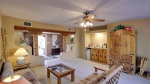 A living room with a sofa, coffee table, and a ceiling fan. There is a kitchen area with wooden cabinets and a brick wall. - Tucson Home - Hiking Trail Access On-site! (Tucson)