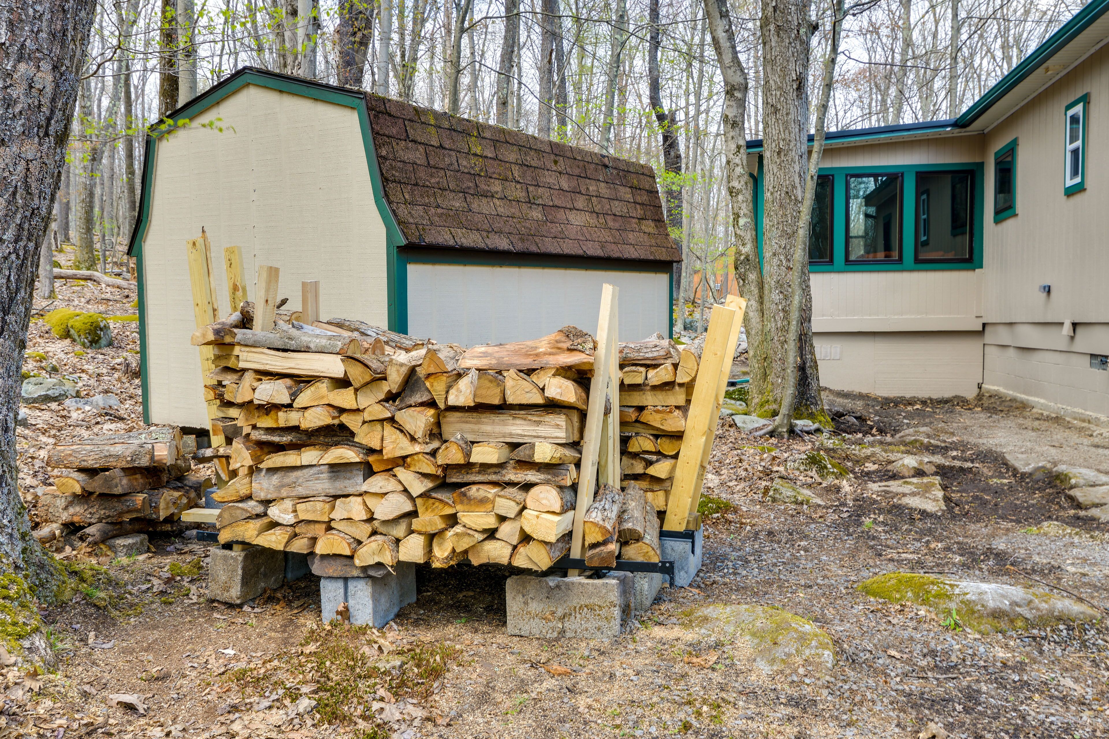 A wooden shed with a stack of firewood in front of a house with a green roof and windows.