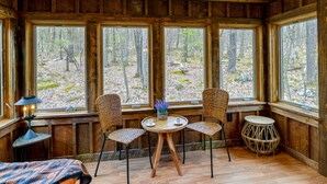 A cozy cabin interior with wooden walls, large windows, a round wooden table, two wicker chairs, and a small round table with a lamp. - Alpine Lake Resort Cabin Rental w/ Pool Access! (Terra Alta)