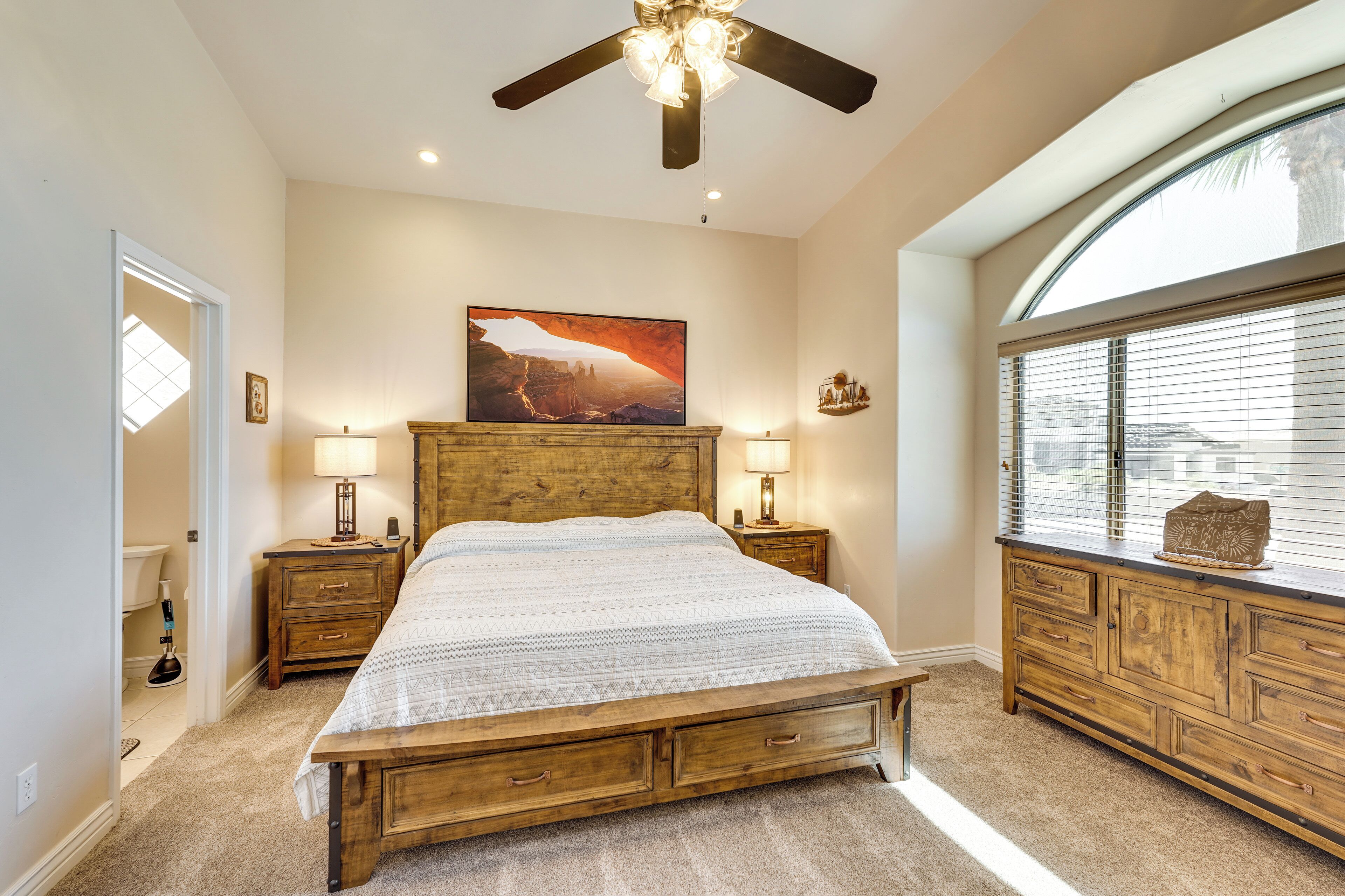 A bedroom with a wooden bed, two bedside tables with lamps, a large window with blinds, and a ceiling fan.