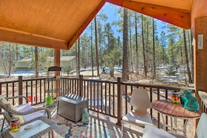 A wooden deck with wicker chairs and a table, overlooking a forested area with tall trees and a clear sky.