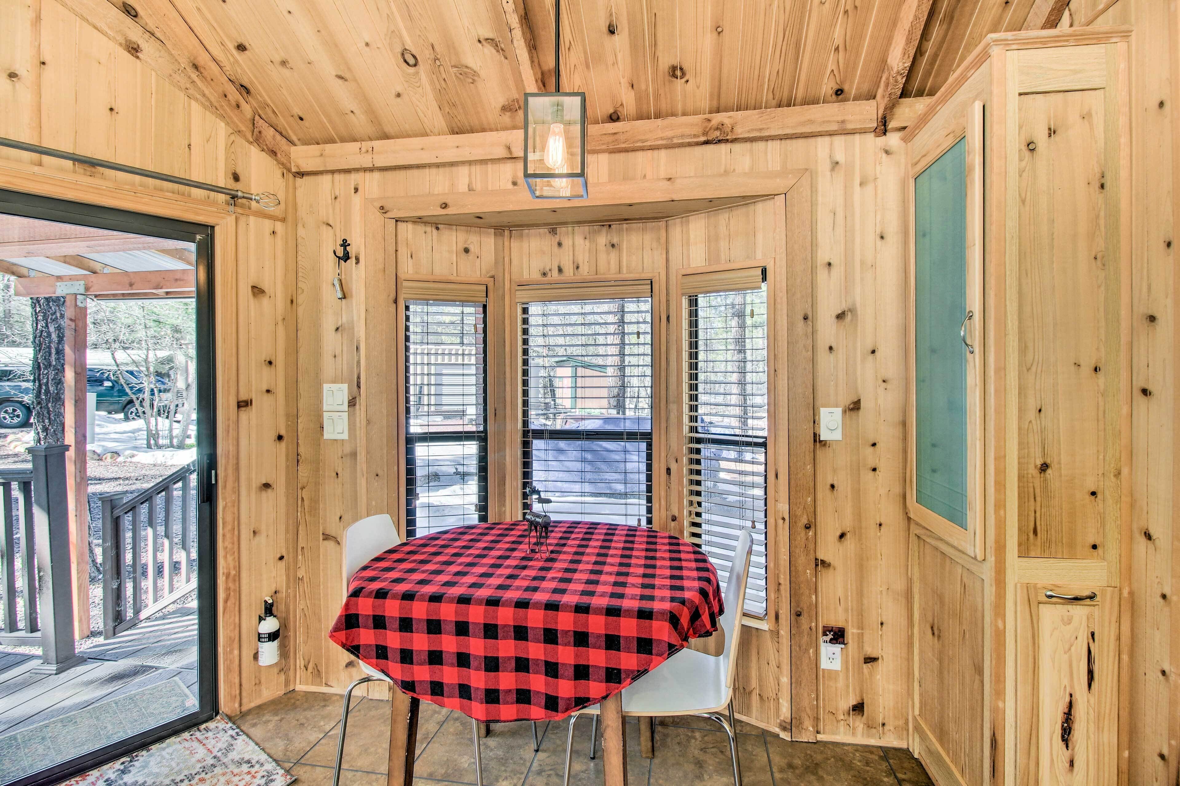 A wooden interior with a round table and two chairs, large windows, and a door.