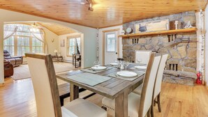 Un coin repas avec une table en bois dressée pour quatre personnes, une cheminée en pierre et une vue sur l’extérieur.