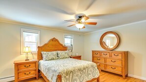 Une chambre à coucher avec un lit en bois, des tables de chevet, une commode, un ventilateur de plafond et deux fenêtres.