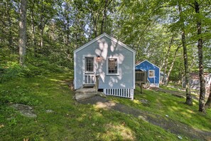 A small, single-story cabin with a white exterior and a blue door, surrounded by a wooded area. - Lush Edgecomb Studio Cottage w/ Fire Table! (Edgecomb)