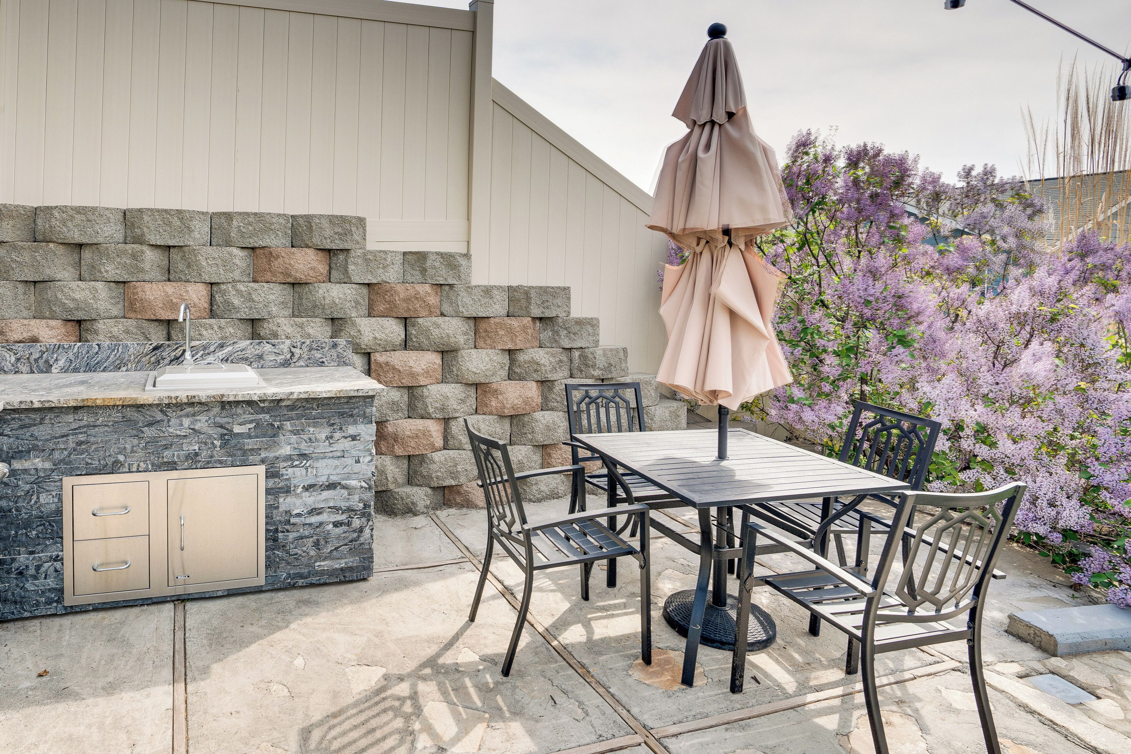 Una terrazza esterna con un muro in pietra, un barbecue integrato, un tavolo con sedie e un cespuglio fiorito di colore viola sullo sfondo.