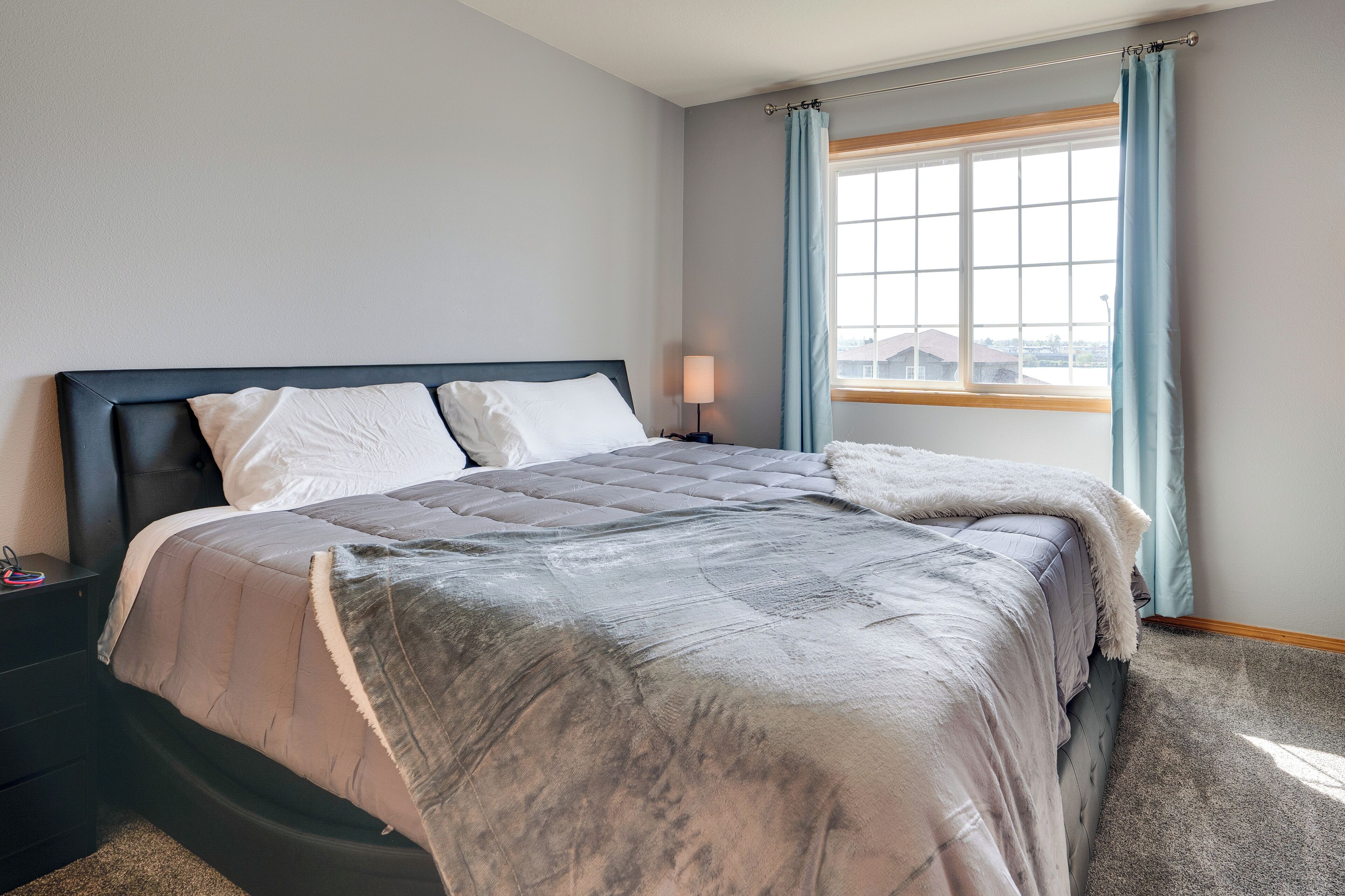 A neatly made bed with a grey duvet and white pillows, a bedside table with a lamp, and a window with light blue curtains.