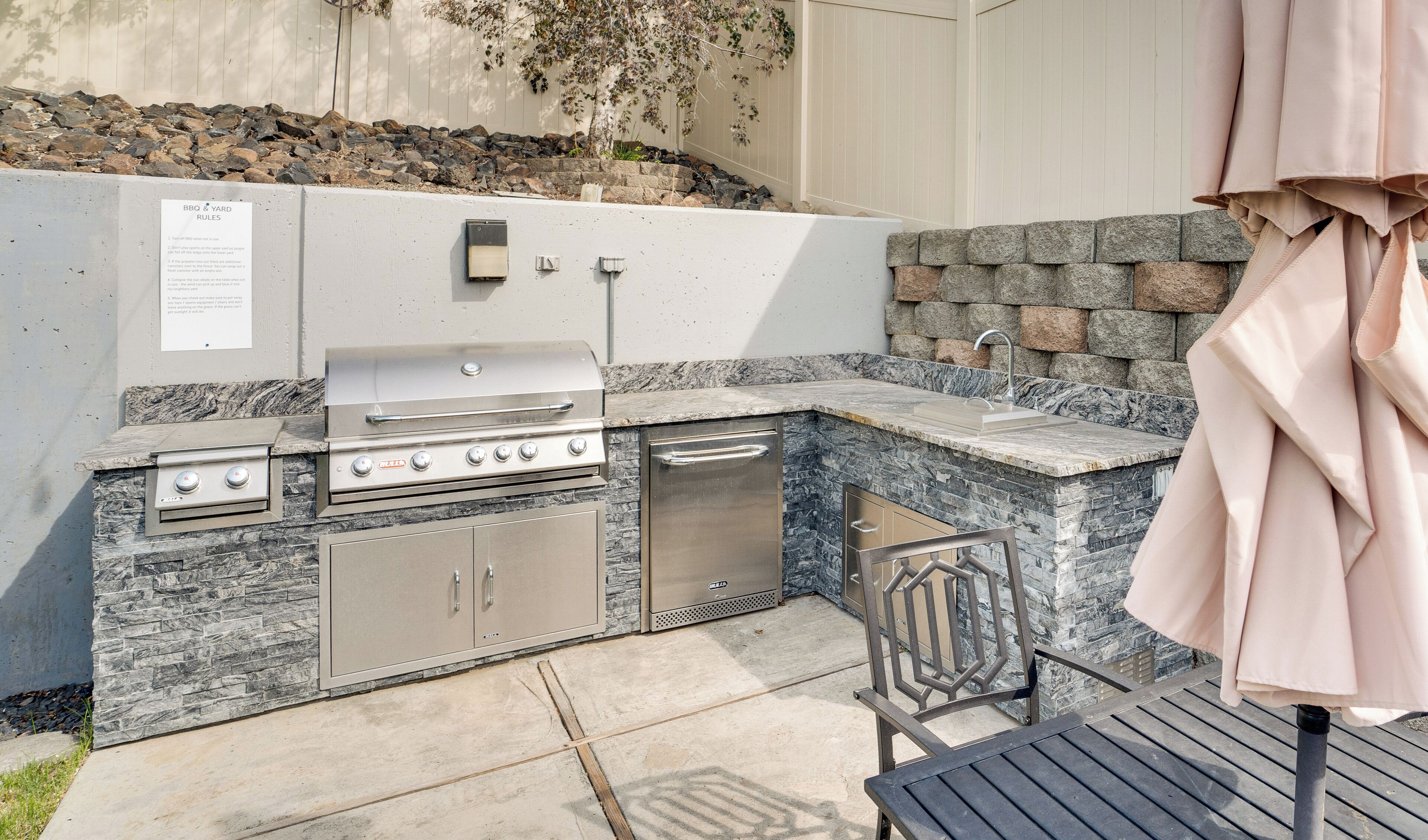 An outdoor kitchen with a grill, sink, and storage cabinets, set against a stone wall with a rock garden.