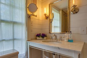 Une salle de bain avec un lavabo en marbre blanc, un miroir rond, une douchette à main et un arrangement floral posé sur le comptoir.