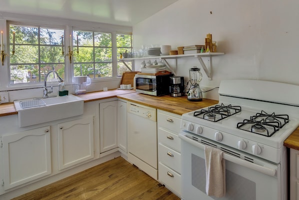Una cocina con armarios blancos, encimera de madera, un fregadero, una estufa y una ventana con vistas a árboles.