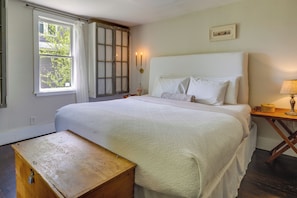Un dormitorio con una cama grande, un cofre de madera, una ventana con cortinas y una lámpara fijada a la pared.