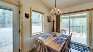 Un comedor con mesa de madera, sillas, una araña y ventanas grandes con vistas a árboles.
