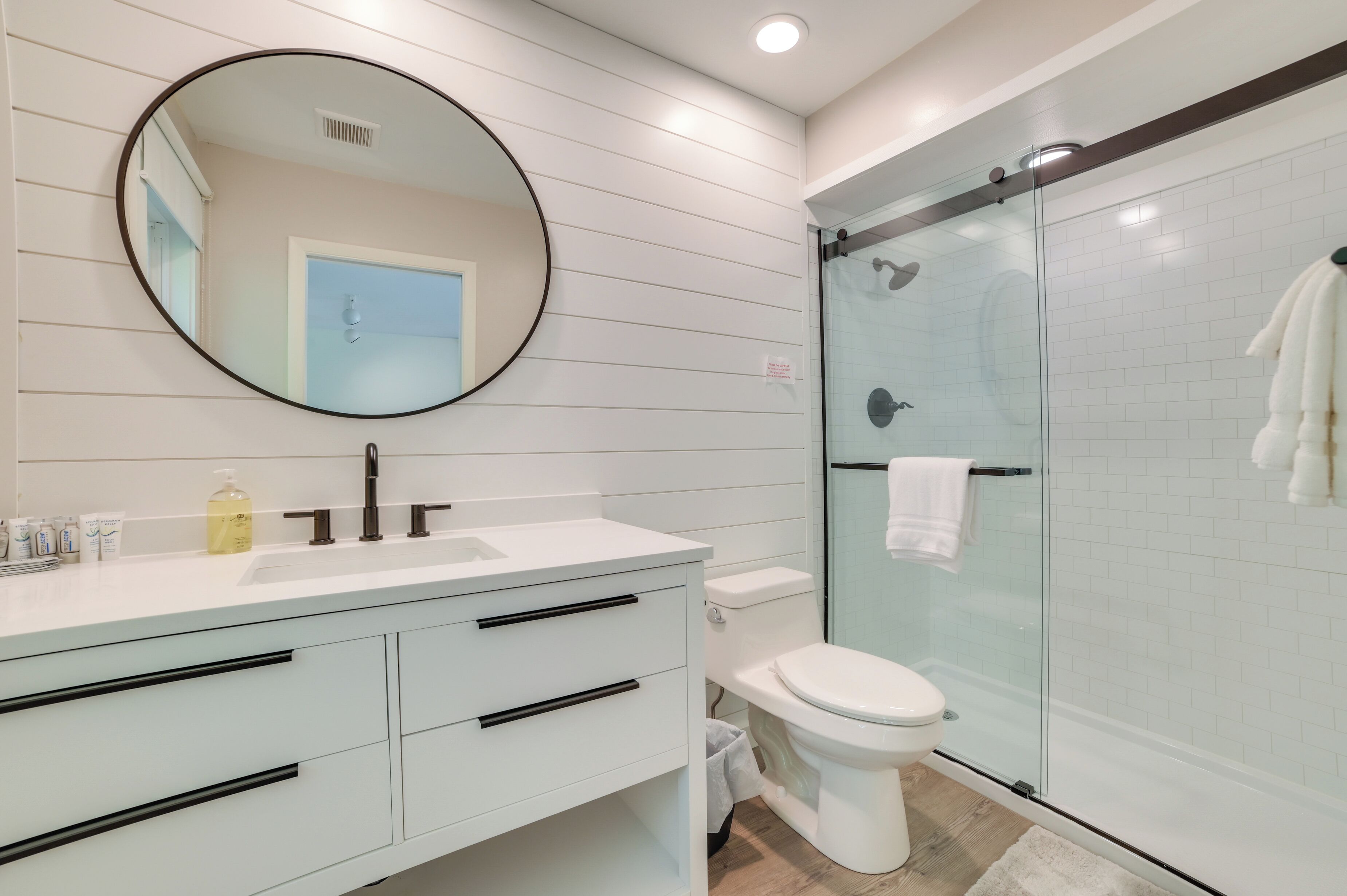 A modern bathroom with a white vanity, a round mirror, a glass shower enclosure, and a white toilet.