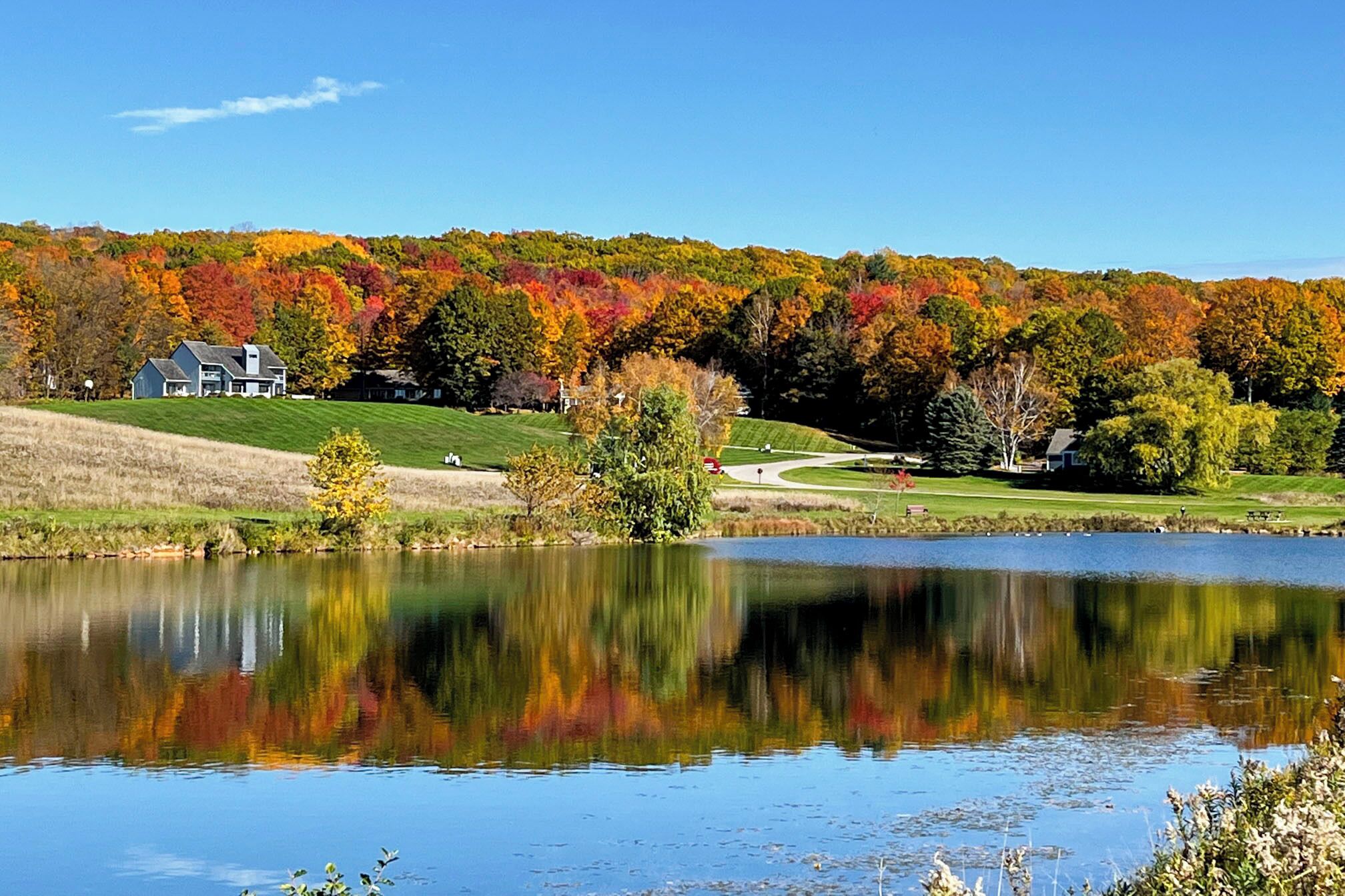 A lake with a reflection of trees, a house on a hill, and a golf course in the background.