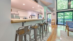 A modern kitchen with a bar area, wooden bar stools, and a view of the outdoors through large windows. - 10 Mi to Nubs Nob: Modern Retreat w/ Fire Pit (Harbor Springs)