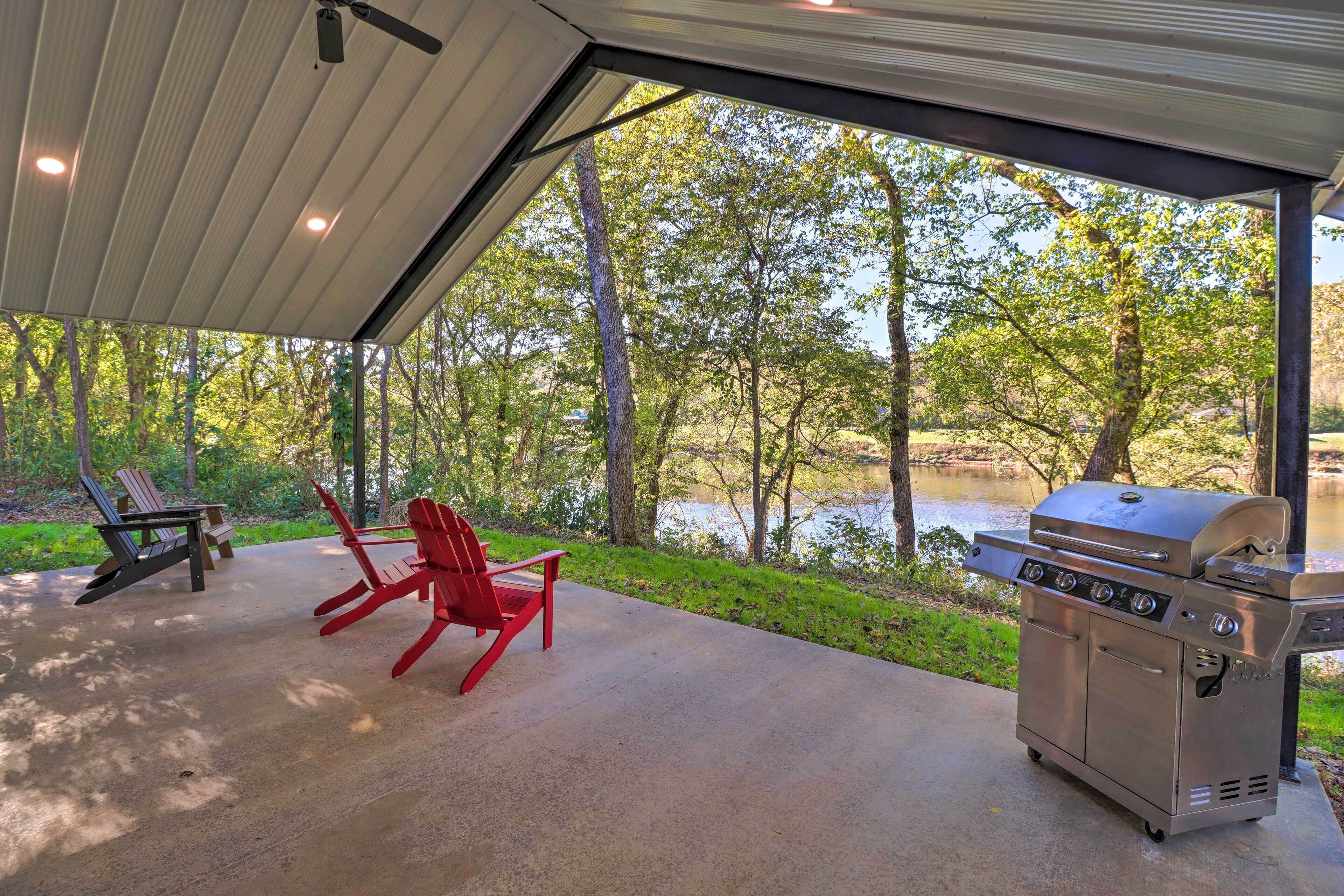 An outdoor covered patio with a grill, red Adirondack chairs, and a view of a lake surrounded by trees.