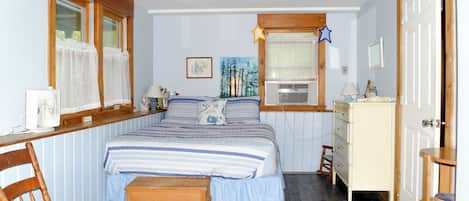 Een slaapkamer met een bed, een houten stoel, een dressoir, een raam met gordijnen en een schilderij aan de muur.
