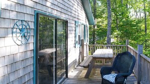 A wooden deck with a glass door, a wicker chair, and a wooden table. - Cozy Waterfront Cottage on Montsweag Bay! (Wiscasset)