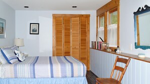 A bedroom with a bed, a wooden chair, a bedside table with books and a lamp, and a window with curtains. - Cozy Waterfront Cottage on Montsweag Bay! (Wiscasset)
