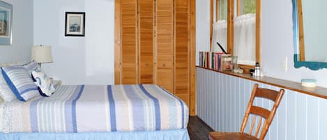Une chambre à coucher avec un lit, une chaise en bois, une table de chevet avec des livres et une lampe, et une fenêtre avec des rideaux.