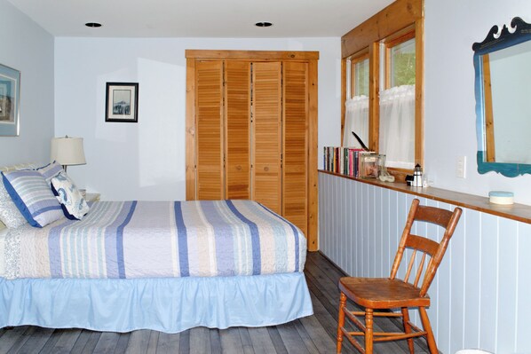 Une chambre à coucher avec un lit, une chaise en bois, une table de chevet avec des livres et une lampe, et une fenêtre avec des rideaux.