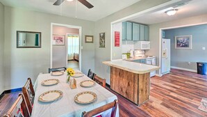 Un coin repas avec une table en bois dressée pour quatre personnes, un ventilateur de plafond et un espace cuisine équipé de placards et d’appareils électroménagers.