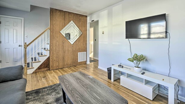 A modern living room with a wooden staircase, a flat-screen TV mounted on the wall, a white console table with a plant, and a grey sofa. - Middletown Township Home, Close to Beaches! (Leonardo)