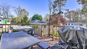 A wooden deck with a table and chairs, a covered grill, and a view of surrounding trees and houses. - Middletown Township Home, Close to Beaches! (Leonardo)