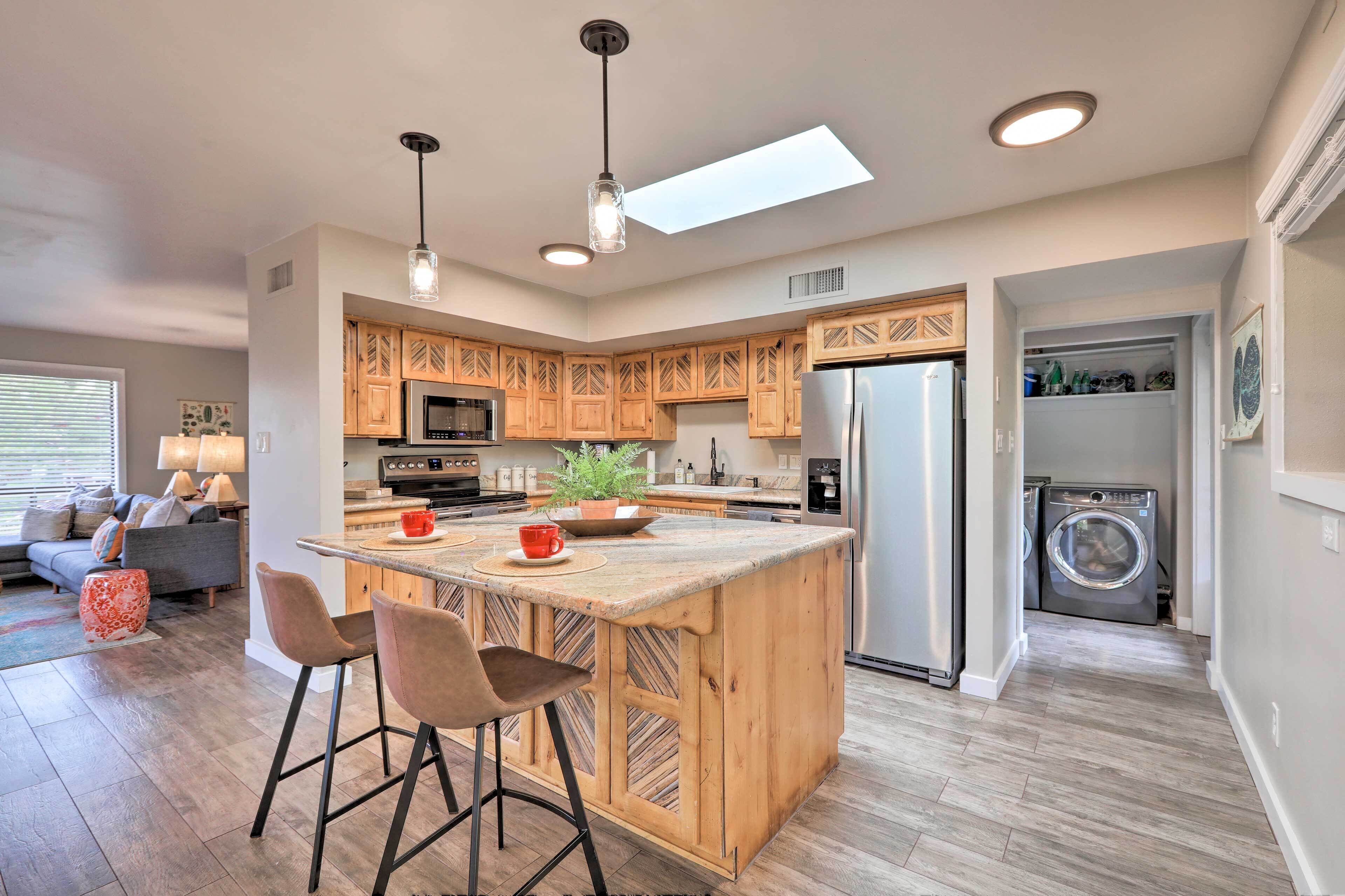 A modern kitchen with wooden cabinets, a central island with bar stools, a built-in oven, and a washing machine in the adjacent room.