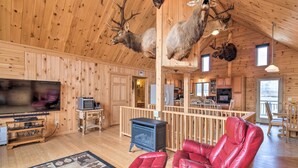 A wooden cabin interior with a fireplace, leather chairs, and mounted animal heads on the ceiling.