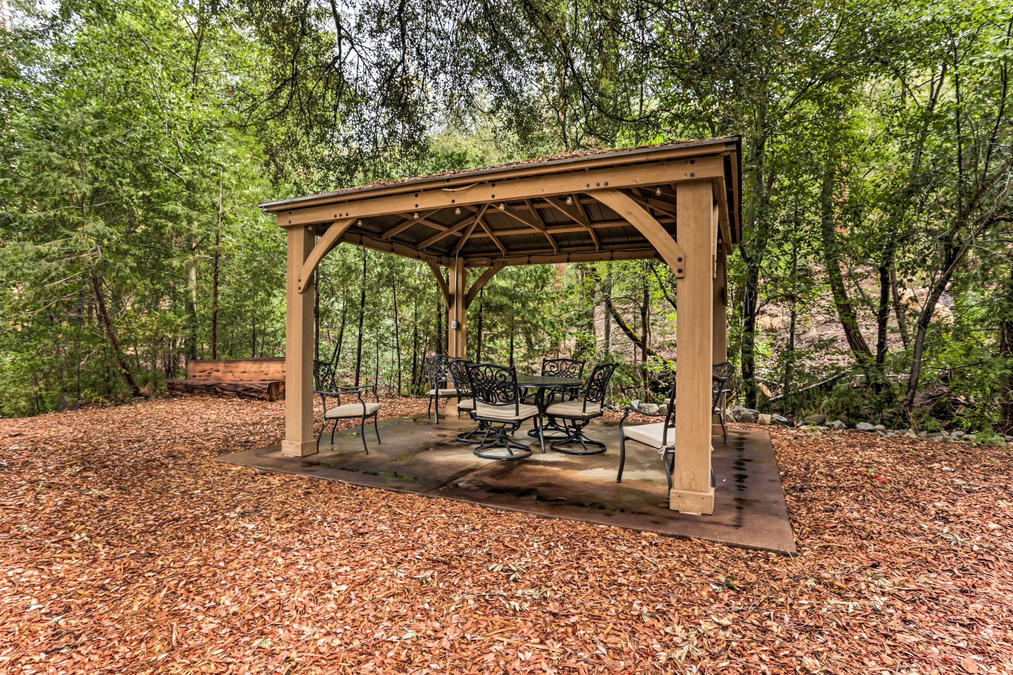A wooden gazebo with a table and chairs in a forested area.
