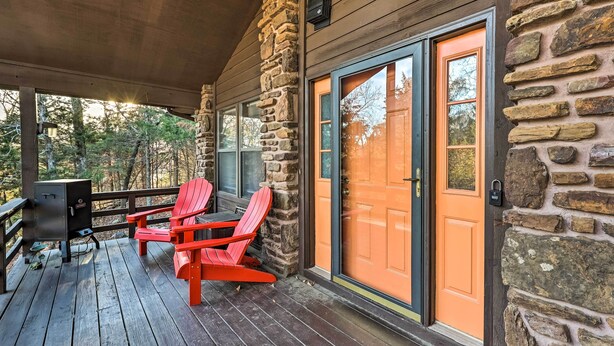 A wooden deck with a stone wall, a sliding glass door, and two red Adirondack chairs. - Cozy Holiday Island Home: Hot Tub + Chiminea! (Eureka Springs)