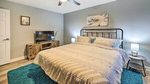 A bedroom with a bed, a ceiling fan, a TV, a small table with a lamp, and a painting on the wall. - Cozy Holiday Island Home: Hot Tub + Chiminea! (Eureka Springs)
