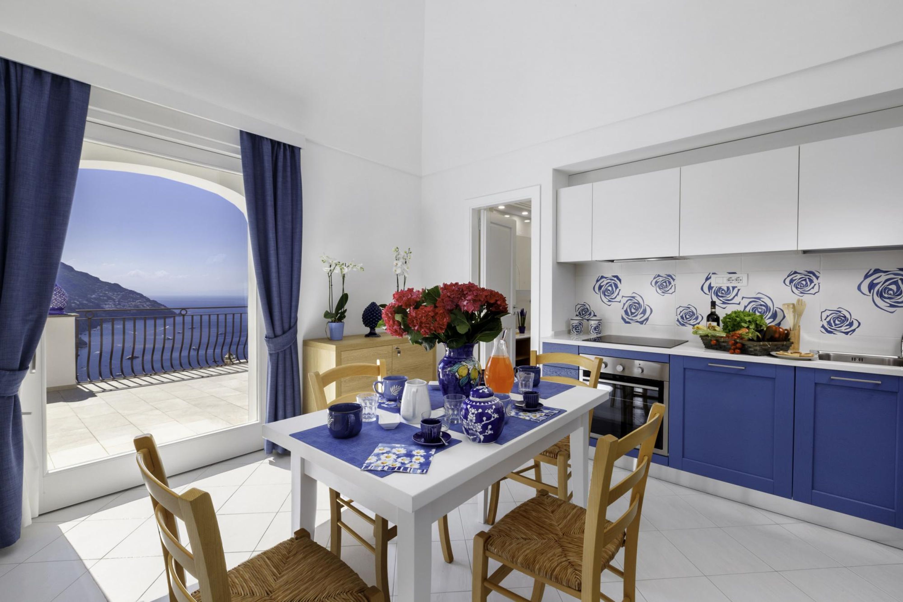 Cuina moderna amb armaris blaus i encimera blanca, zona de dinar amb taula blanca i calessals blaus, i balcó amb vista al mar.